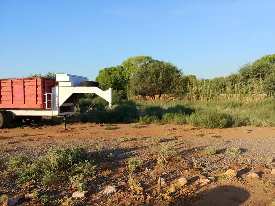 Red and white trailer parked on a dirt lot, surrounded by green shrubs and trees, under a blue sky.