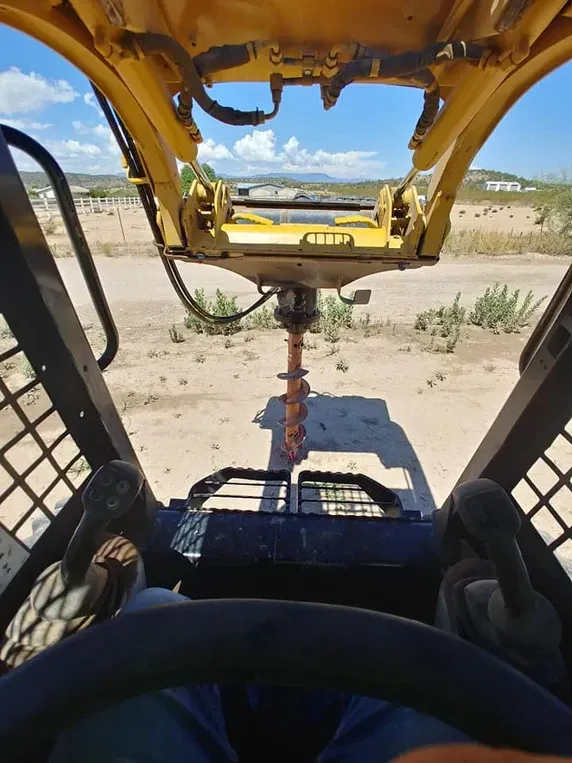 View from inside a skid steer operating an auger, on a dirt lot with blue sky.