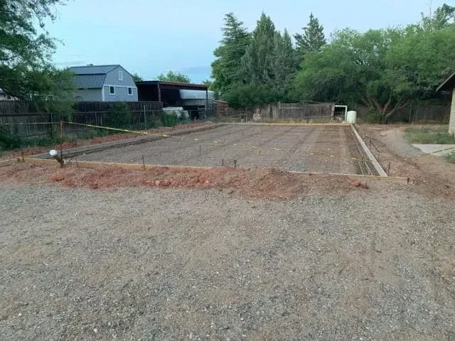 Gravel yard with a rectangular concrete slab under construction, bordered by dirt. A shed and house are in the background.