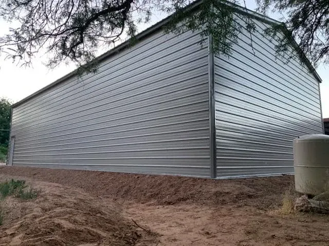 Large, silver metal building under a tree, set on a dirt foundation.