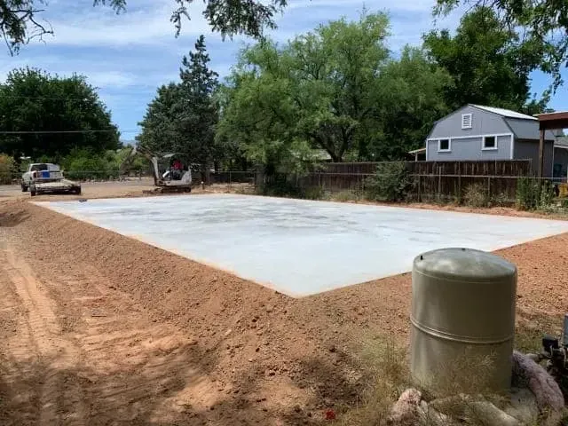 Newly poured concrete slab in dirt yard, construction site. A water tank is in foreground.
