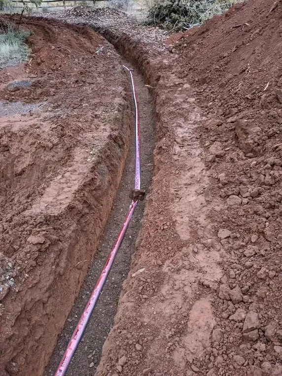Trench dug into reddish soil, with a pink measuring tape running along the bottom.