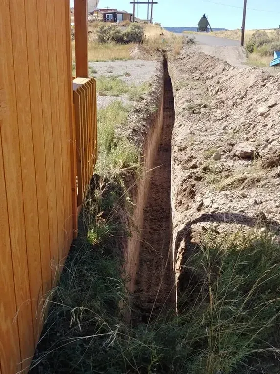 A deep, narrow trench dug alongside a wooden fence, dirt path, and grassy area.