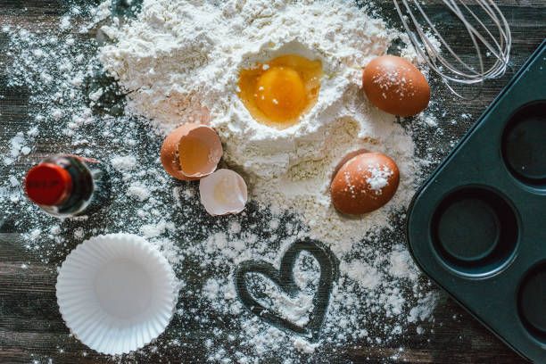 Baking ingredients arranged on a dark surface: flour, eggs, muffin tin, whisk, vanilla, and a heart drawn in flour.