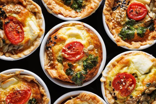 Overhead shot of individual quiches in white ramekins. Each with tomato and other toppings on a dark surface.