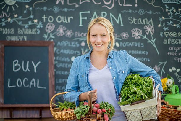 Woman at a farm stand, holding baskets of produce, smiling. 