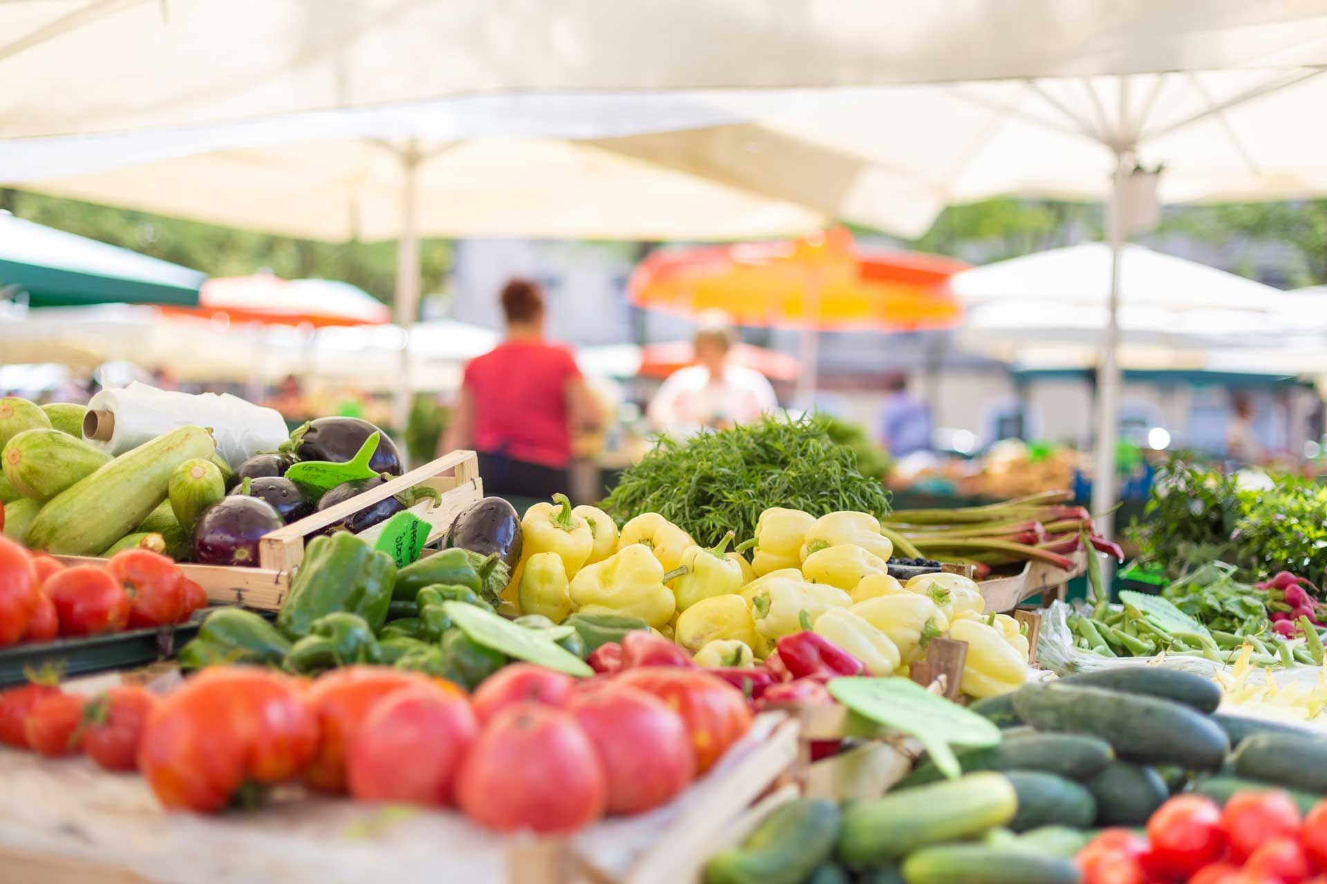 Produce display of fresh vegetables at a farmer's market with customers in the background.
