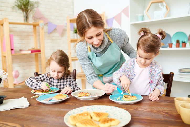 Woman and two young children decorate pancakes at a table; colorful plates, light-filled room.