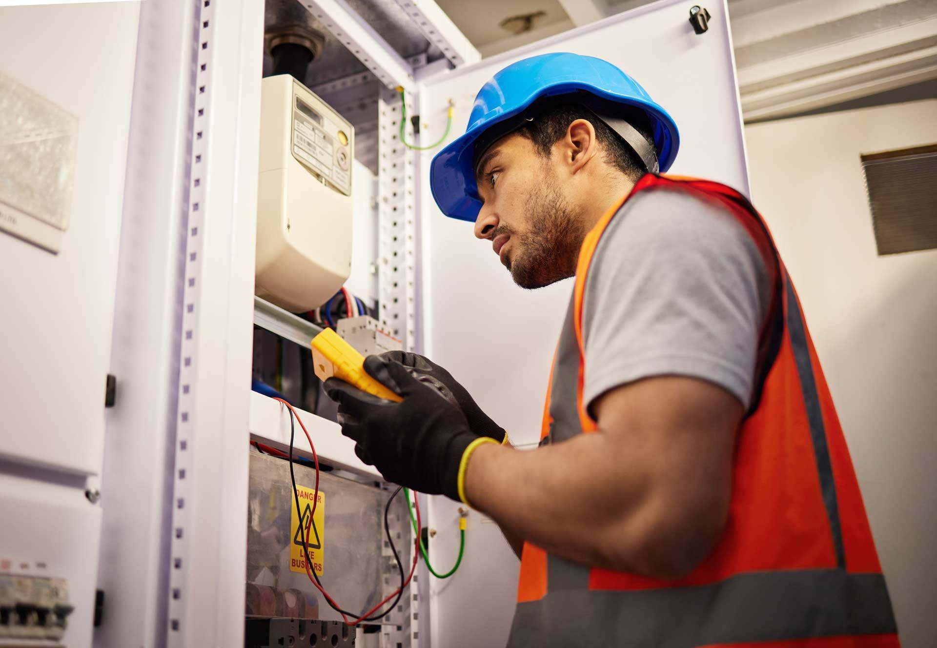 Electrician in safety gear examines electrical panel with a multimeter.