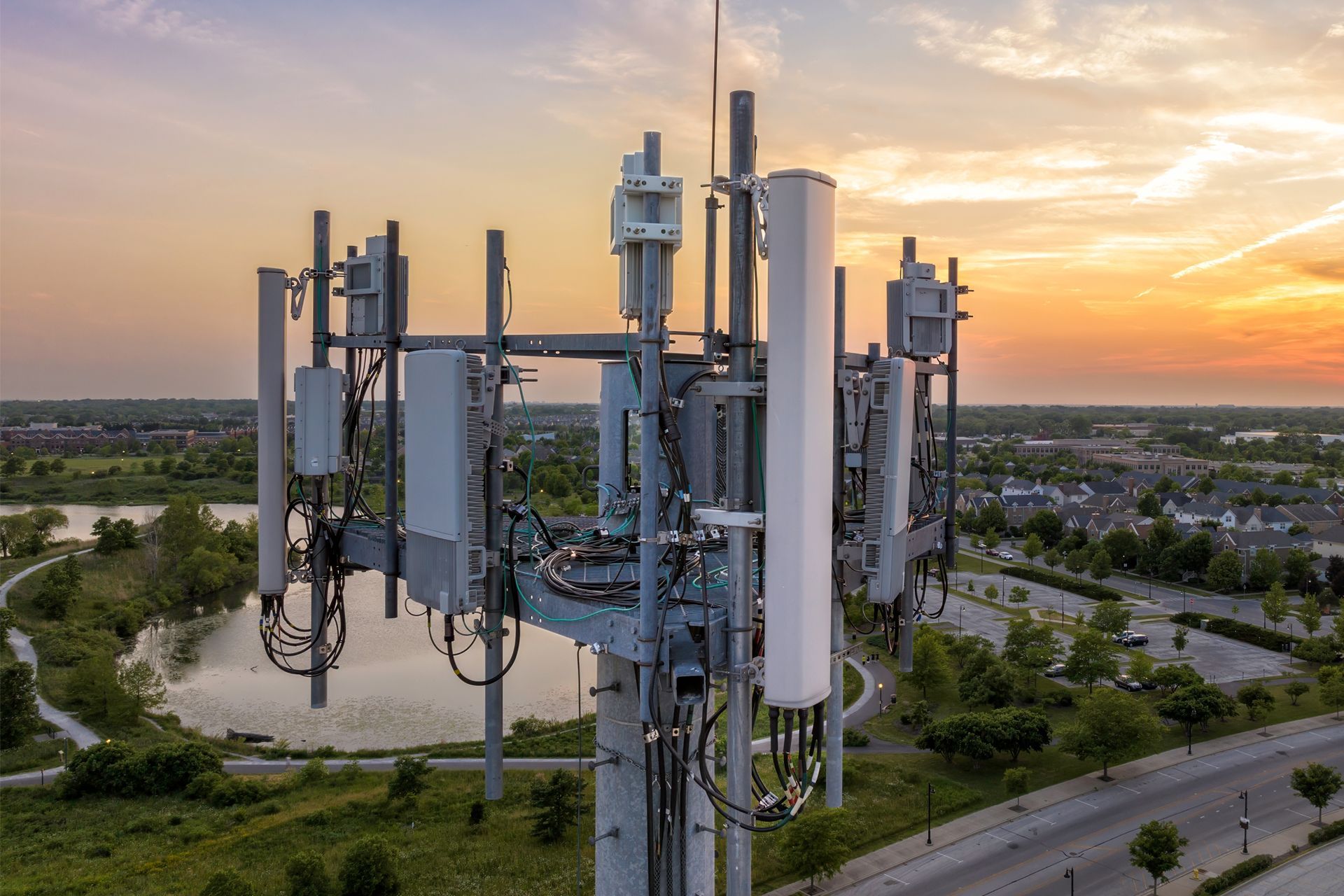 Cell tower with various antennas against a sunset sky, overlooking a park and road.