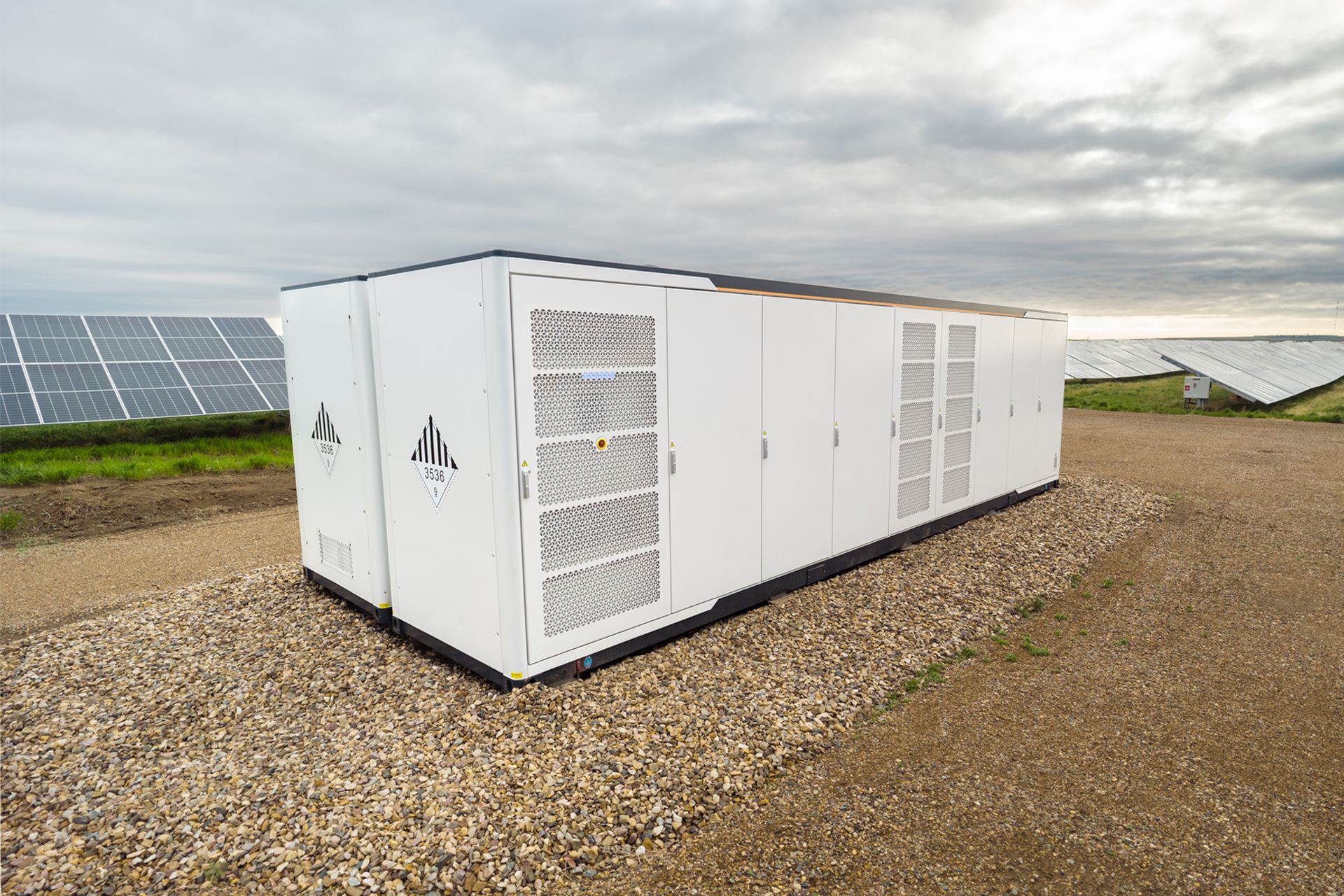 White energy storage unit with vents, in a solar panel field under a cloudy sky.