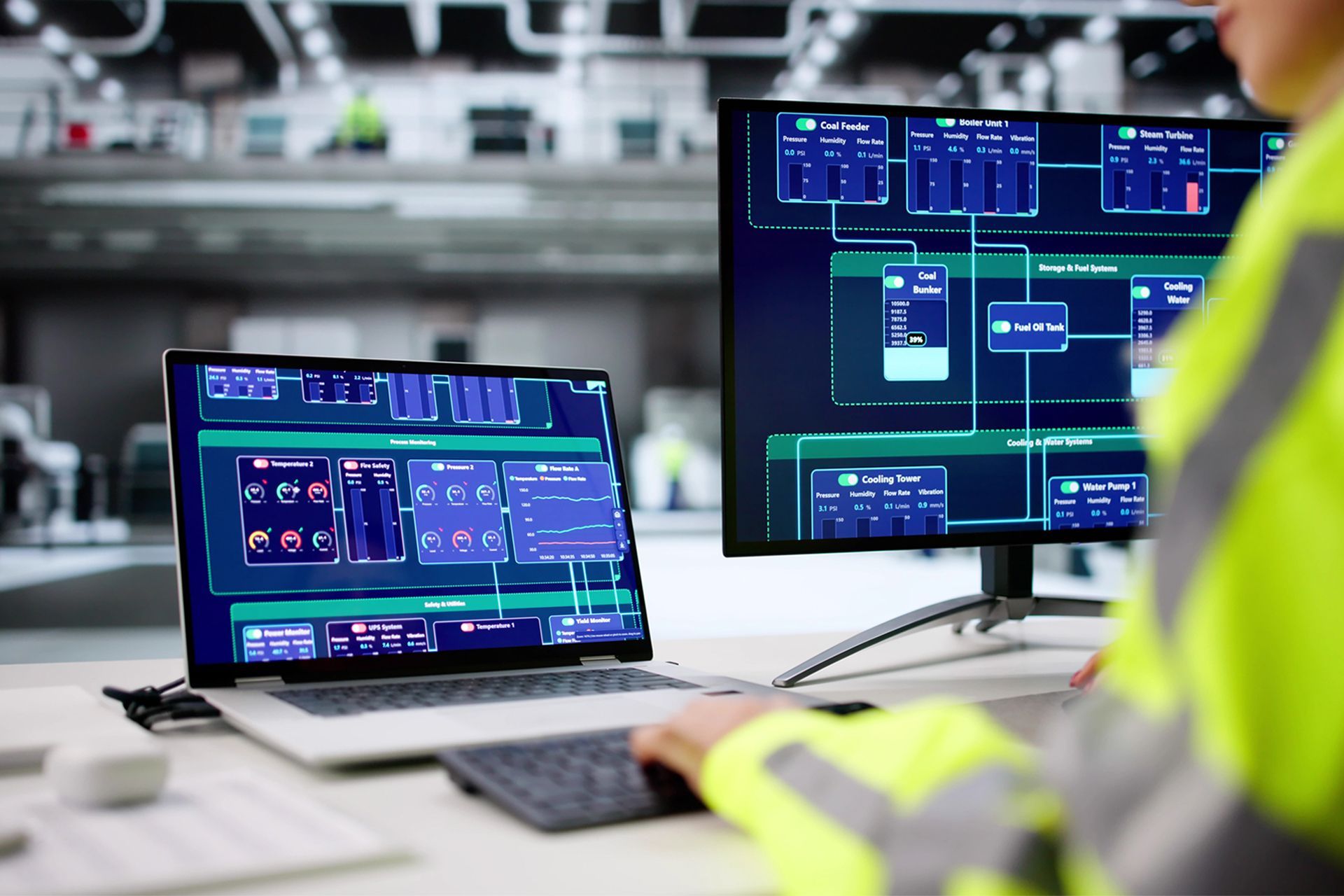 Person in a yellow safety vest monitors data on two computer screens in a factory setting.