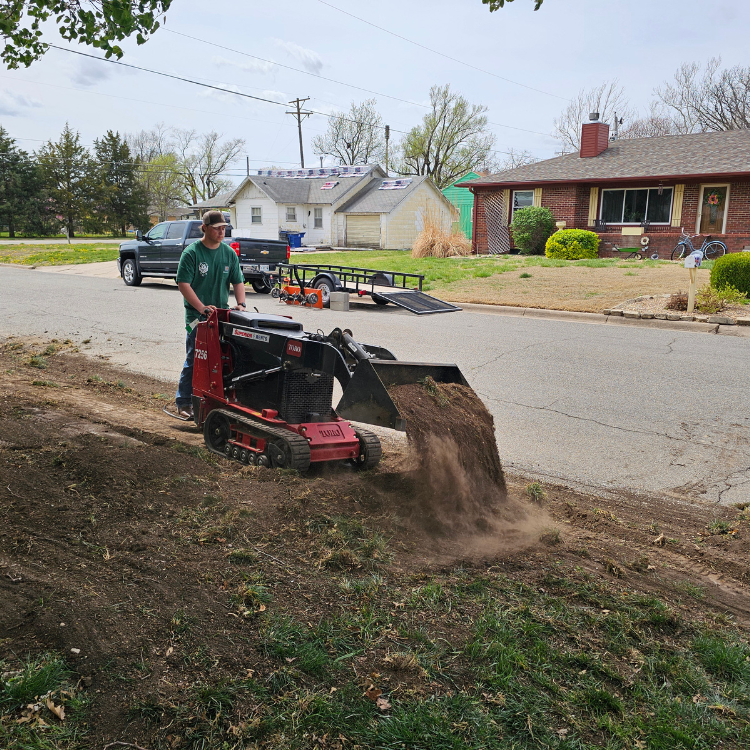 Man operating a red skid steer, dumping dirt onto a grassy slope on a residential street.
