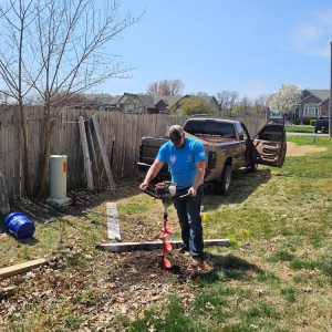 Man using a post hole digger in a backyard, brown truck, wooden fence, blue sky.