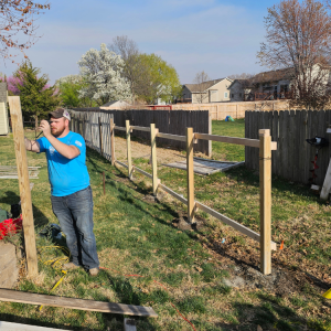 Man in blue shirt builds wooden fence in a grassy backyard on a sunny day.