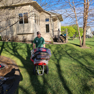 Man in green shirt pushing a wheelbarrow full of supplies across a grassy yard near a house.