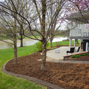 Lush brown mulch bed surrounds trees on a green lawn overlooking a lake and a patio.