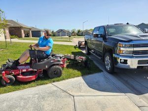Man mowing lawn with a zero-turn mower, truck and trailer parked on side of the road in a suburban neighborhood.