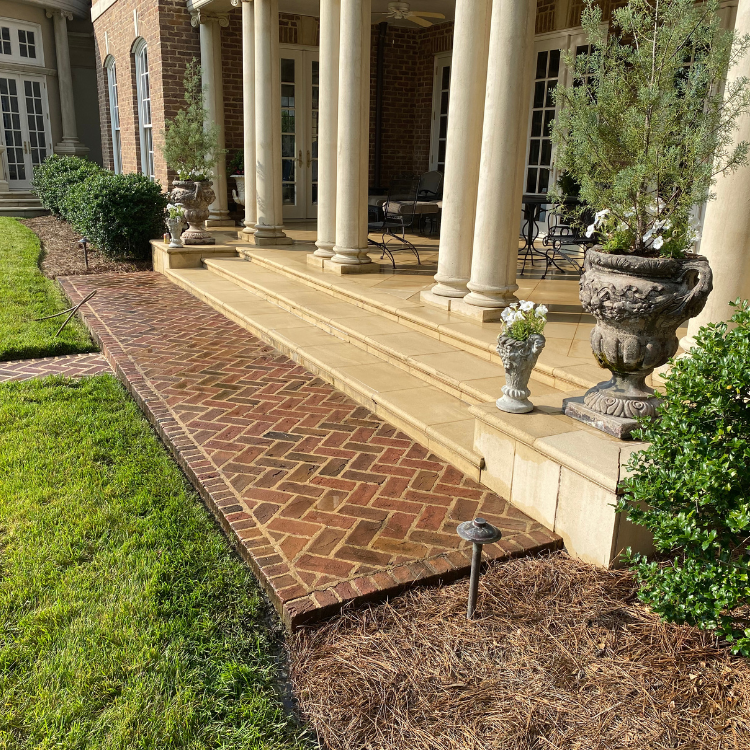 Brick pathway leads to a porch with columns, urns, and landscaping. Green grass is on the left.