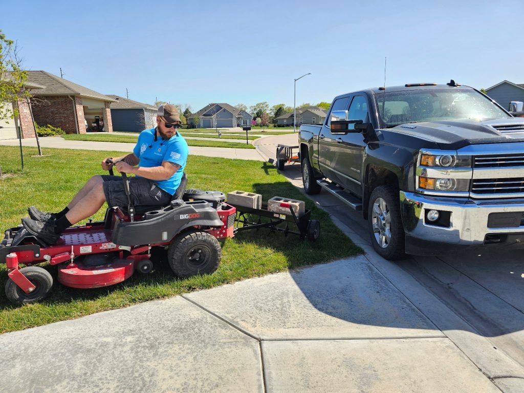Man on a red lawnmower pulls a trailer next to a black truck on a sunny suburban street.