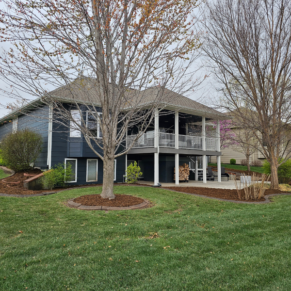 Two-story house with dark blue siding, wraparound deck, and green lawn. Trees and landscaping in front.