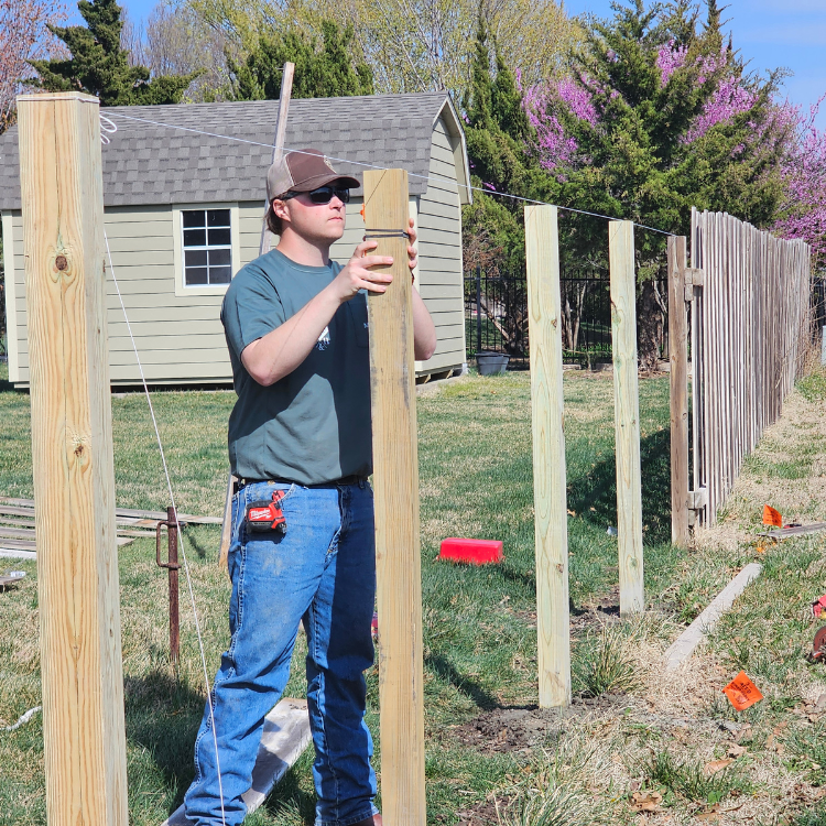 Man building a fence outdoors, holding a wooden post, with a shed and existing fence in the background.