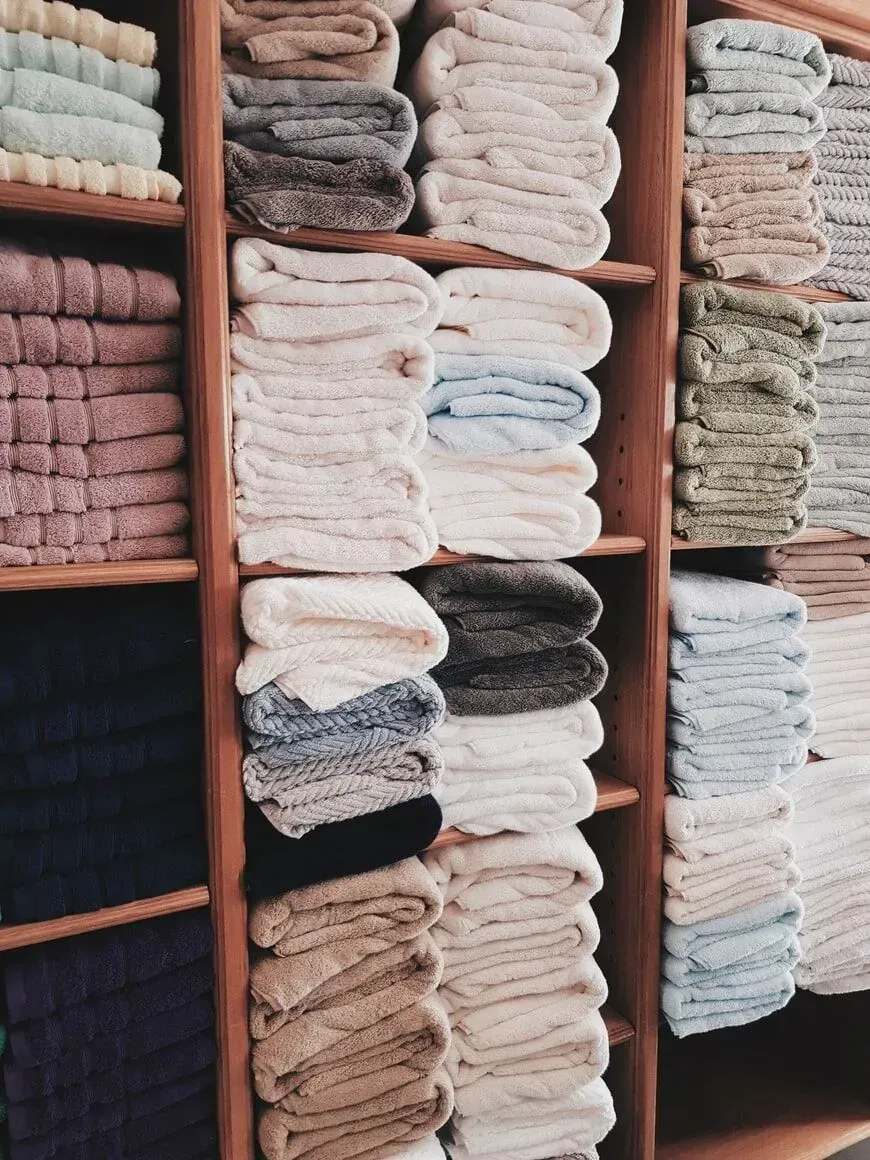 Wooden shelves stocked with neatly folded stacks of towels in various neutral, blue, and pink tones.