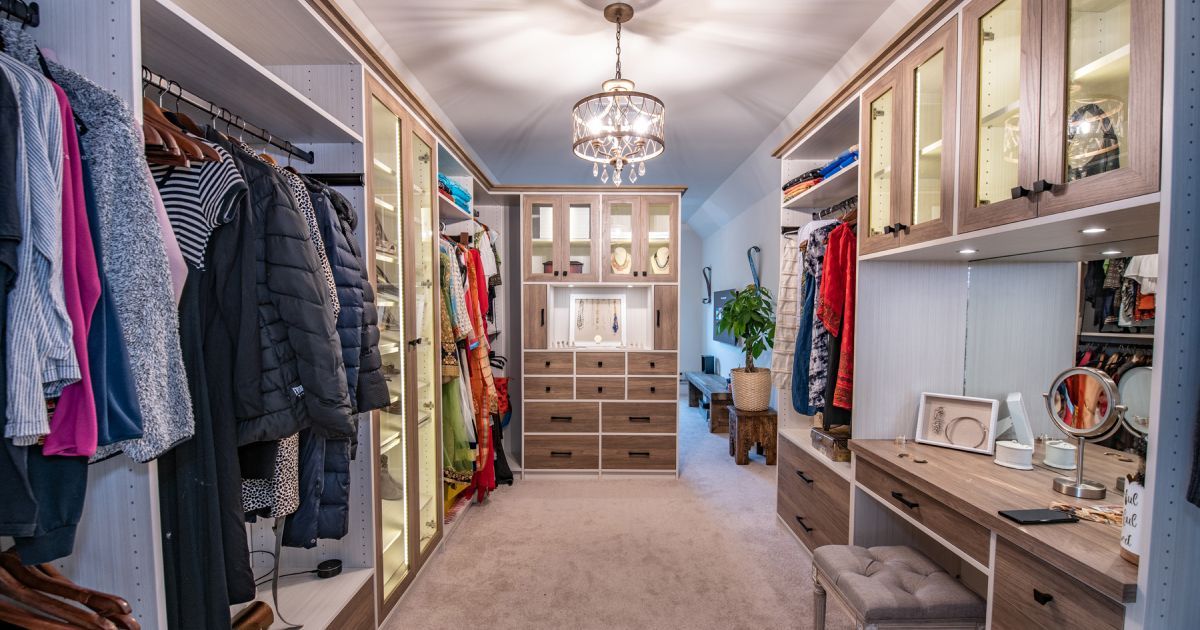 A walk-in closet with hanging clothes, wooden drawers, glass-front cabinets, and a vanity area under a crystal chandelier.