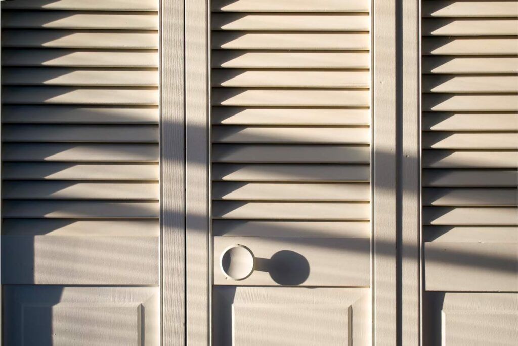 Beige louvered closet doors with shadows cast by sunlight, featuring a circular knob on the center panel.