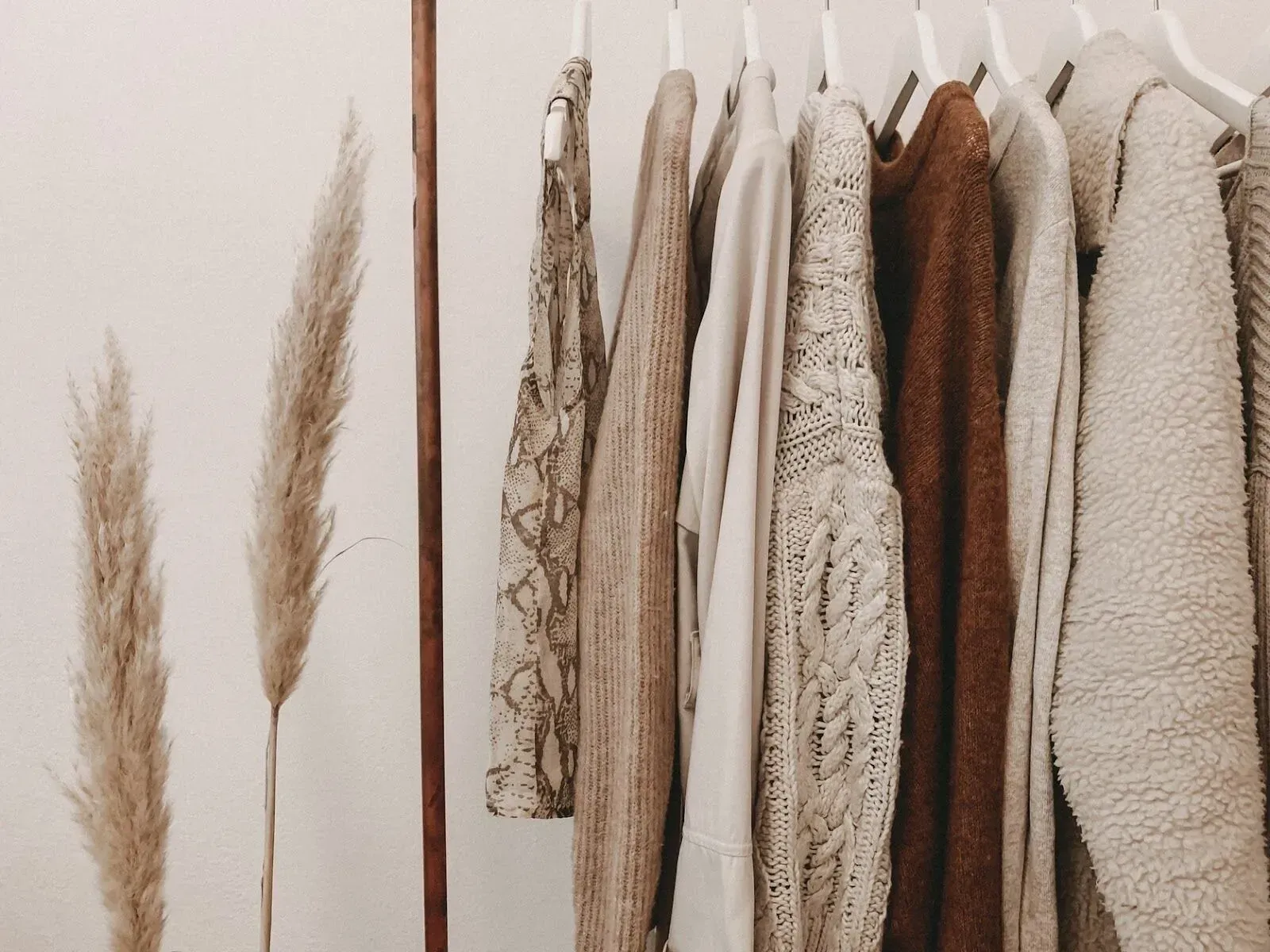 A clothing rack displaying various neutral-toned sweaters and a patterned garment, next to two pampas grass stems.