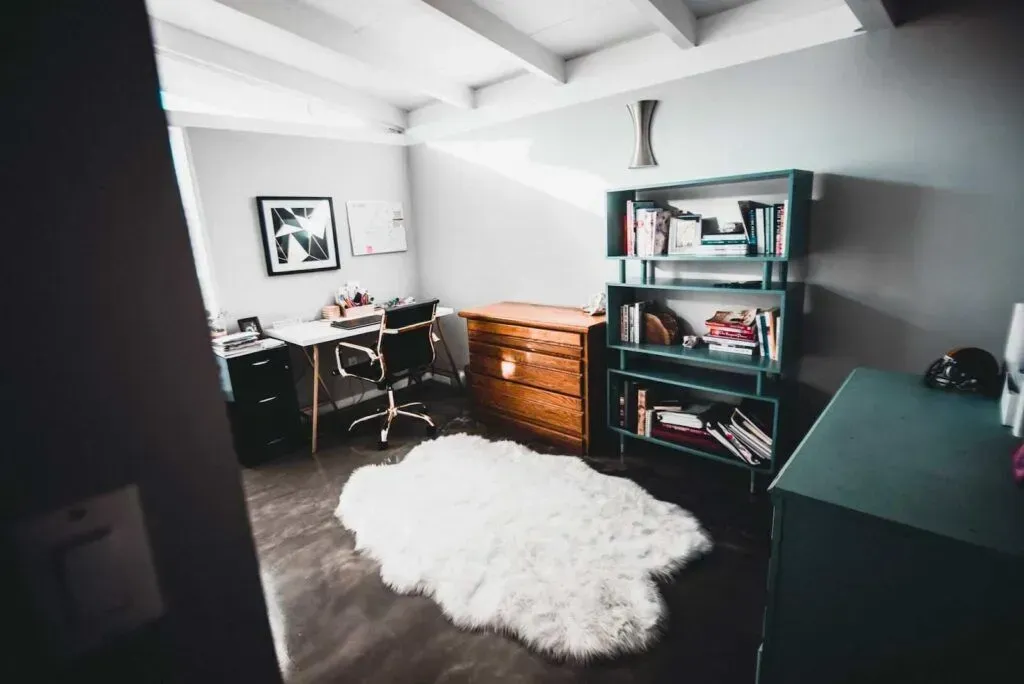 A home office with a desk, swivel chair, wooden chest of drawers, a teal bookshelf, and a white faux-fur rug on the floor.