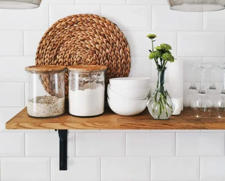 A wooden shelf against a white subway tile wall holds two glass jars, a stack of white bowls, and a vase of green flowers.