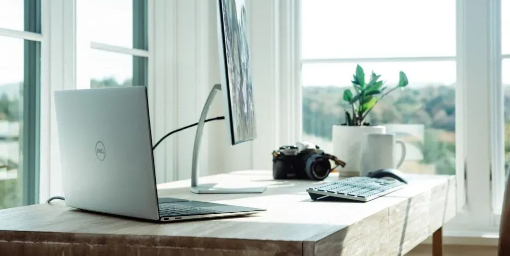 Laptop, monitor, and camera on a desk near a window with trees.