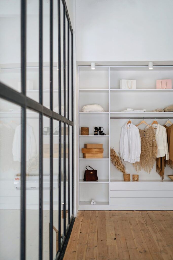 Modern walk-in closet with white shelving, hanging clothes, and wood floors, seen through a black-framed glass partition.