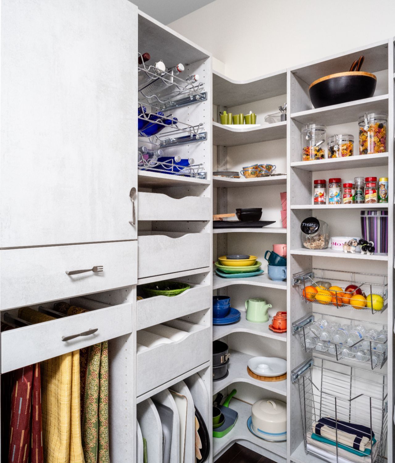 Organized white pantry shelving with various shelves, pull-out drawers, wire baskets, and hanging kitchen linens.