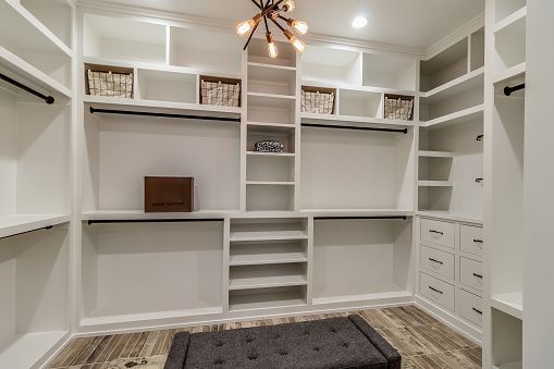 A walk-in closet with white shelving, hanging rods, drawers, and a central tufted bench over a wood-look floor.