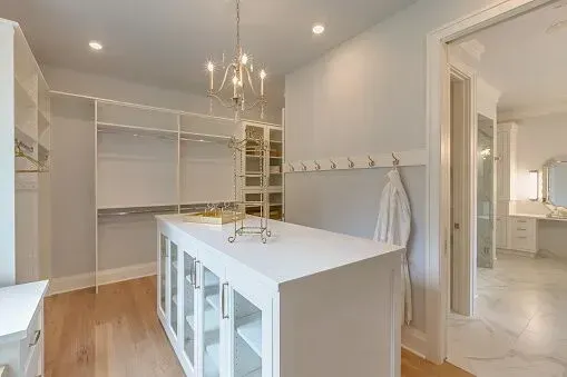 A white walk-in closet featuring a center island, open shelving, a chandelier, and a doorway leading to a bright bathroom.