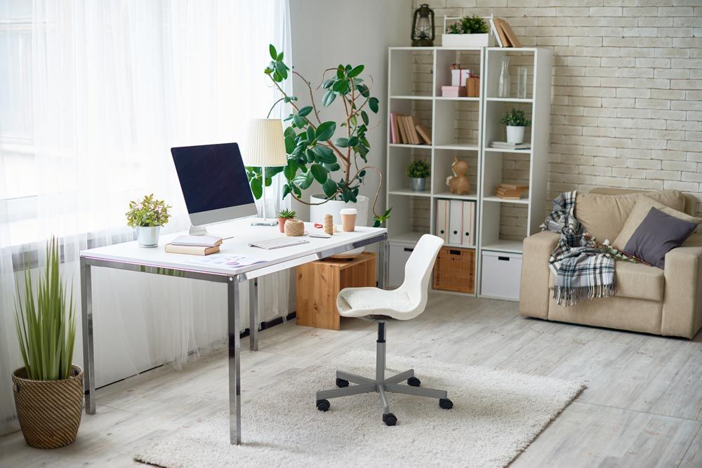 A bright home office with a modern white desk, computer, swivel chair, leafy green plants, and a cozy armchair.