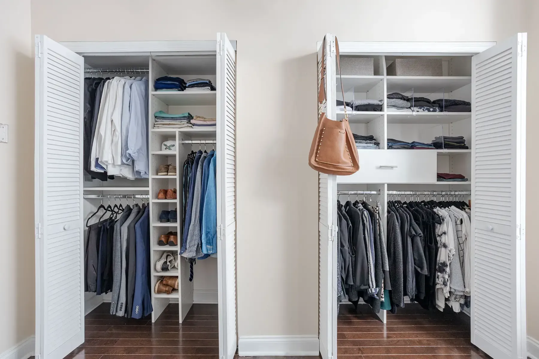 Two organized white reach-in closets with louvered doors, filled with hanging clothes, shelves, and a brown leather bag.