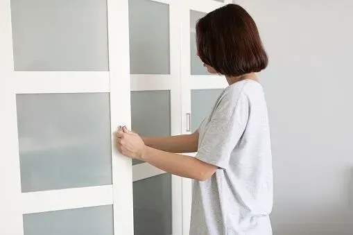 A person in a light grey t-shirt opening a white cabinet with frosted glass panels.