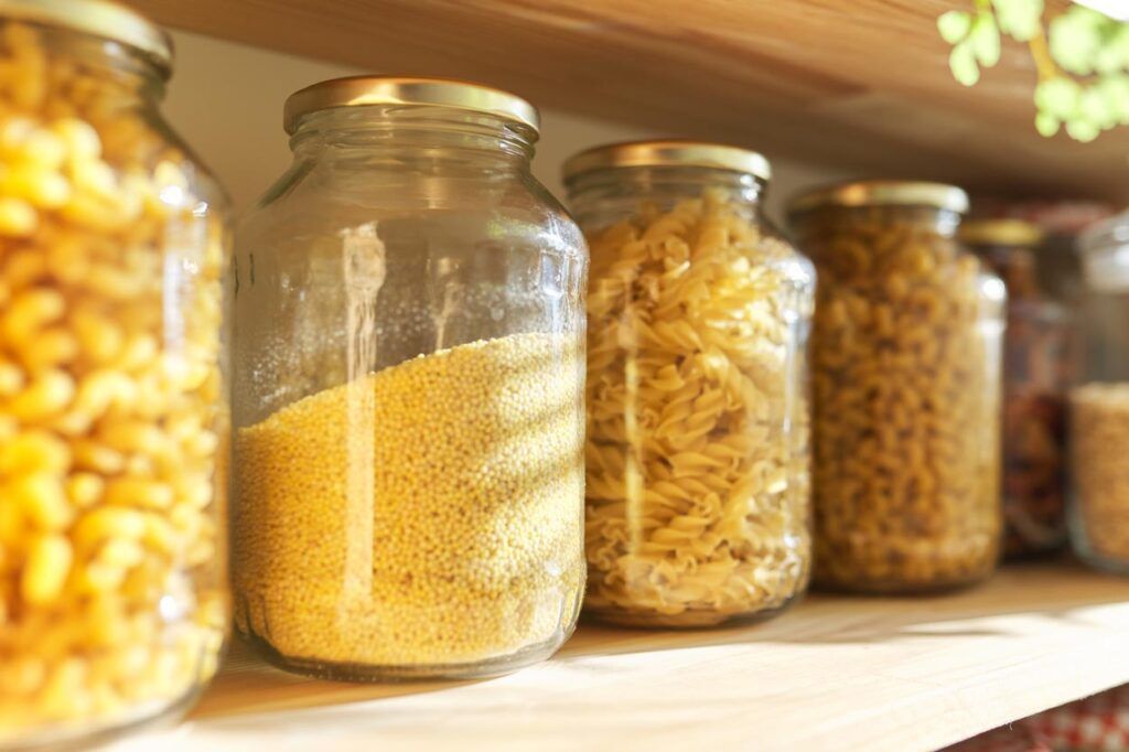 A row of clear glass jars filled with various types of dry pasta and grains, arranged on a wooden kitchen shelf.