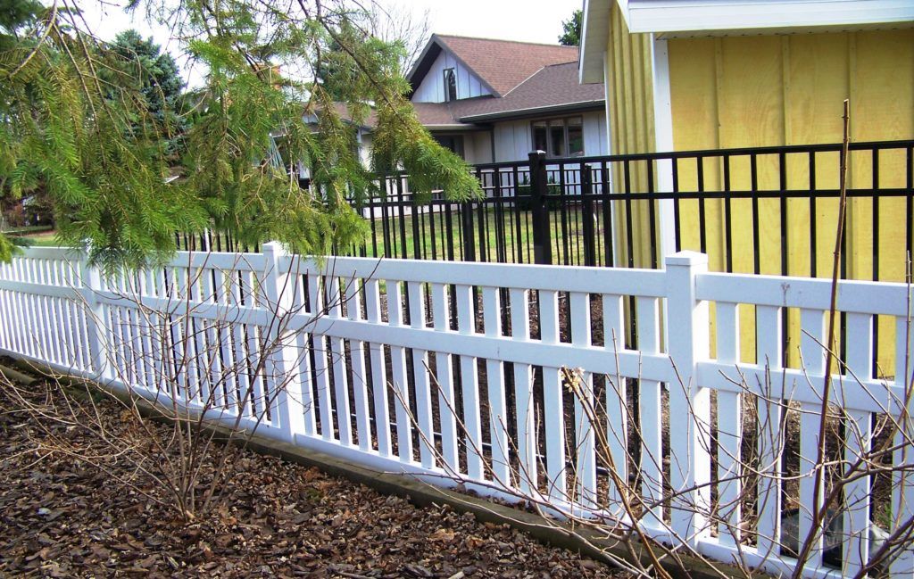 A white vinyl fence with a yellow house & black aluminum fence in the background