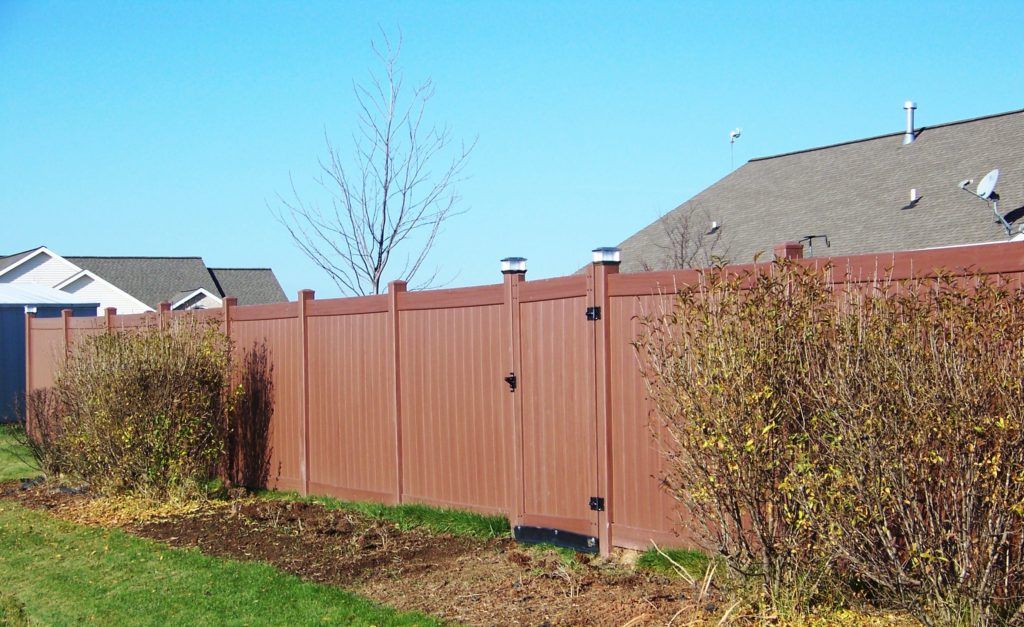 A brown vinyl fence is surrounded by bushes and a house