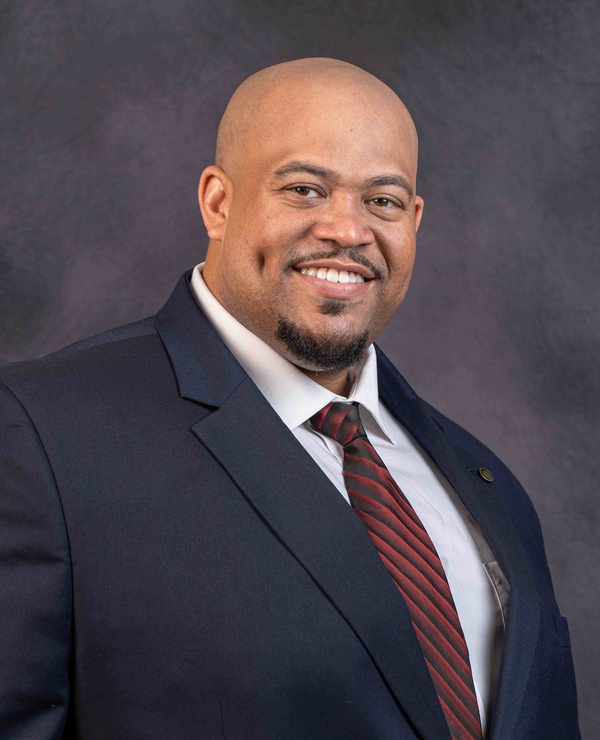 A man wearing a dark blue suit and a maroon striped tie smiles against a mottled gray background.