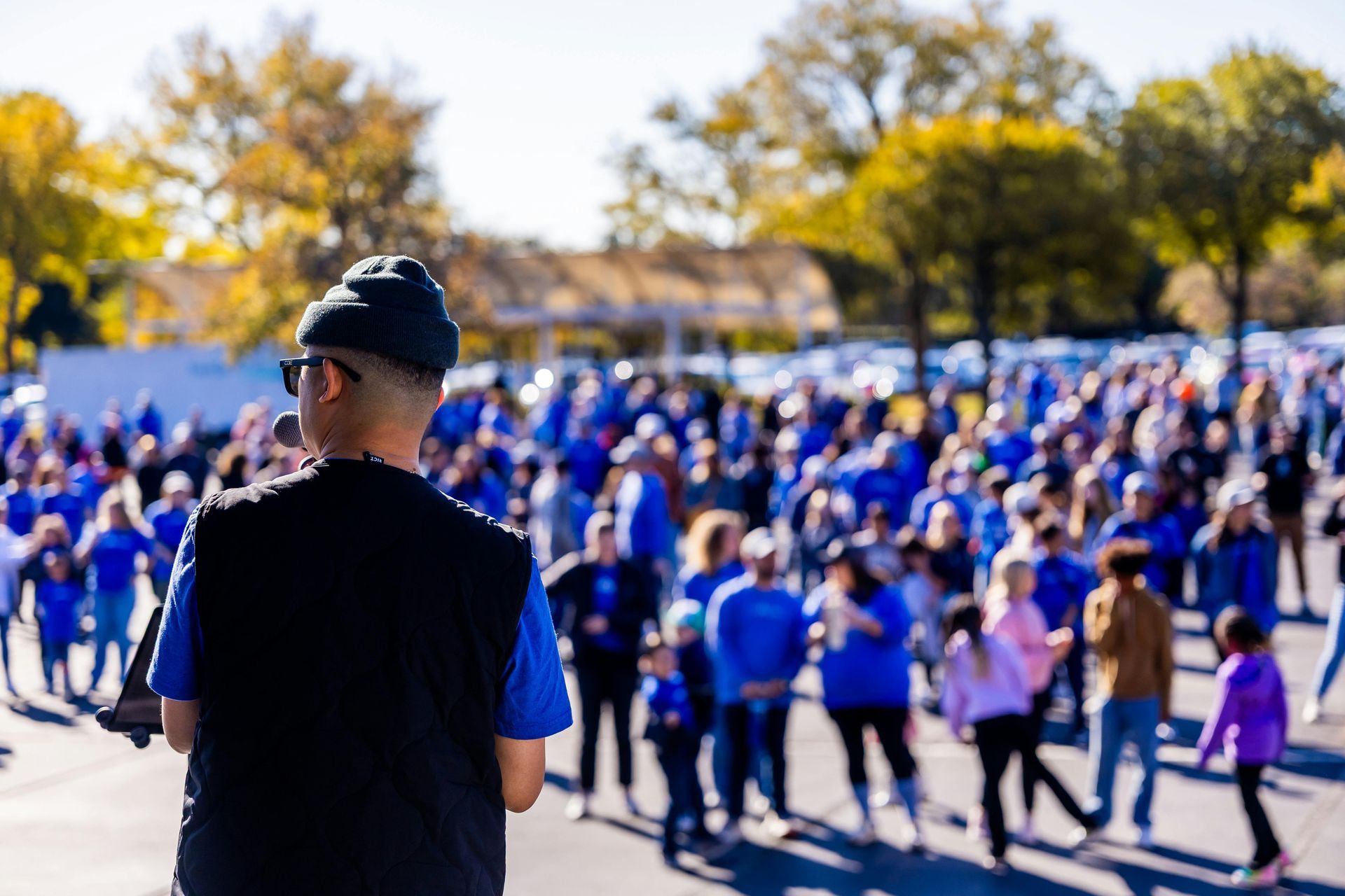 A person in a dark beanie and vest addresses a large, blurred crowd gathered outside on a sunny day.