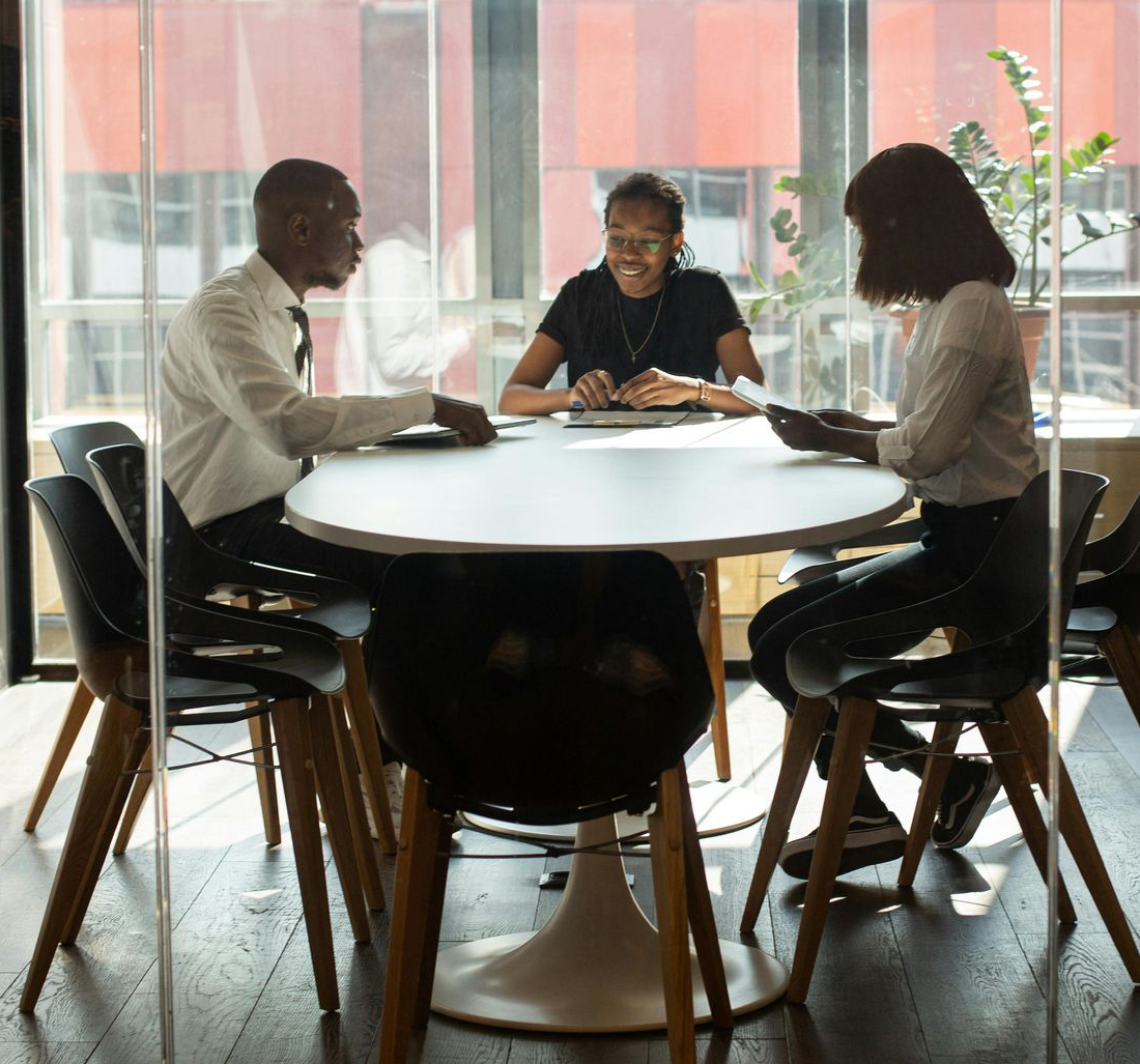Three people in professional attire meet around a round table in a glass-walled conference room.
