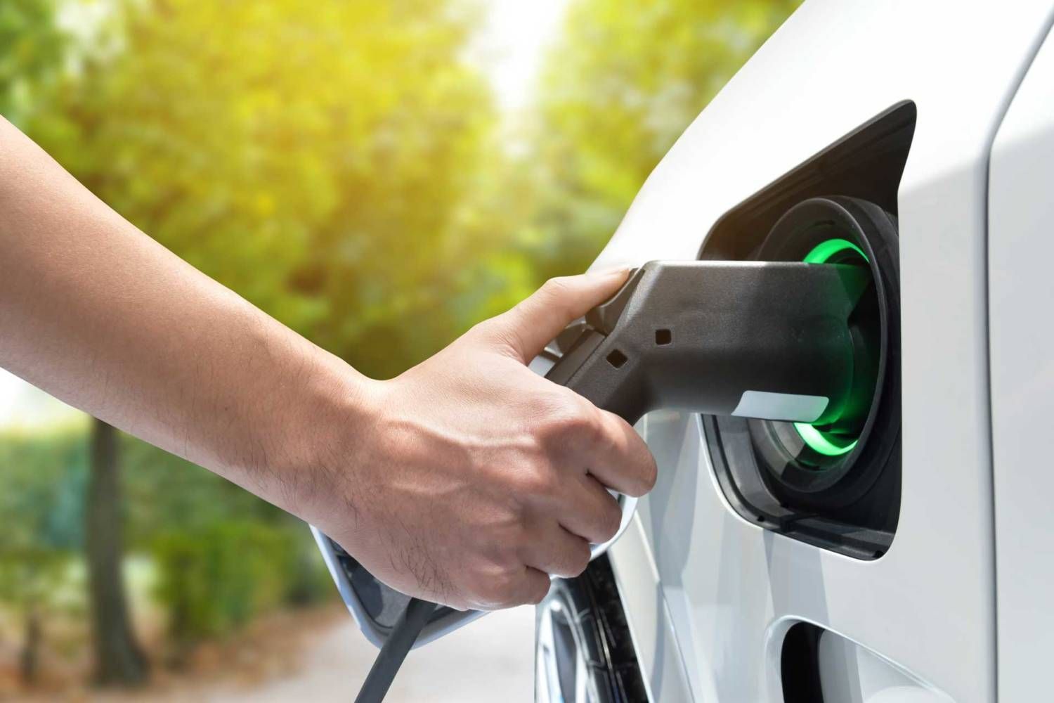 A hand inserts a black charging cable into the port of a white electric vehicle, indicated by a glowing green light.