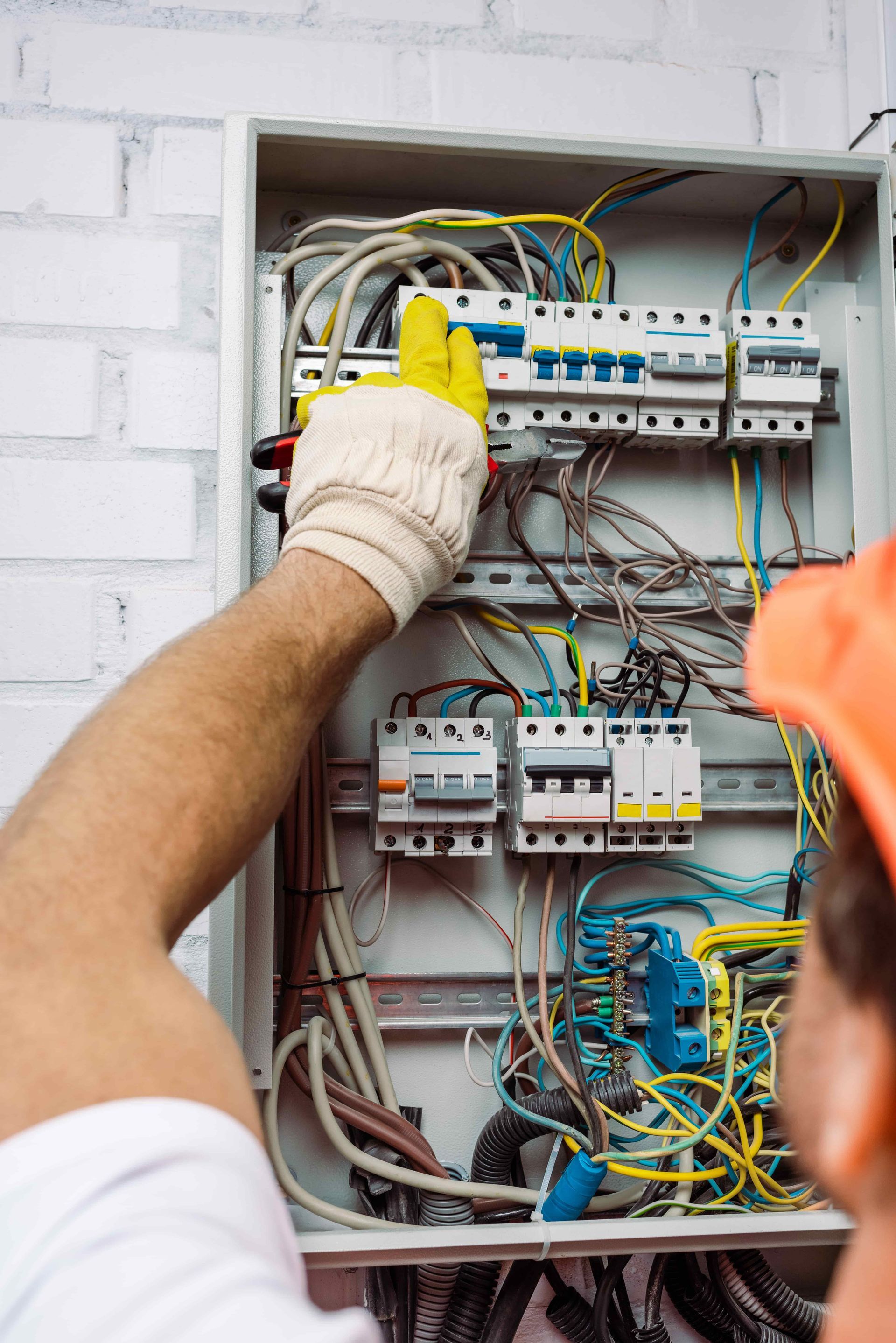 An electrician wearing work gloves and an orange hard hat installs wiring in an open electrical distribution panel.