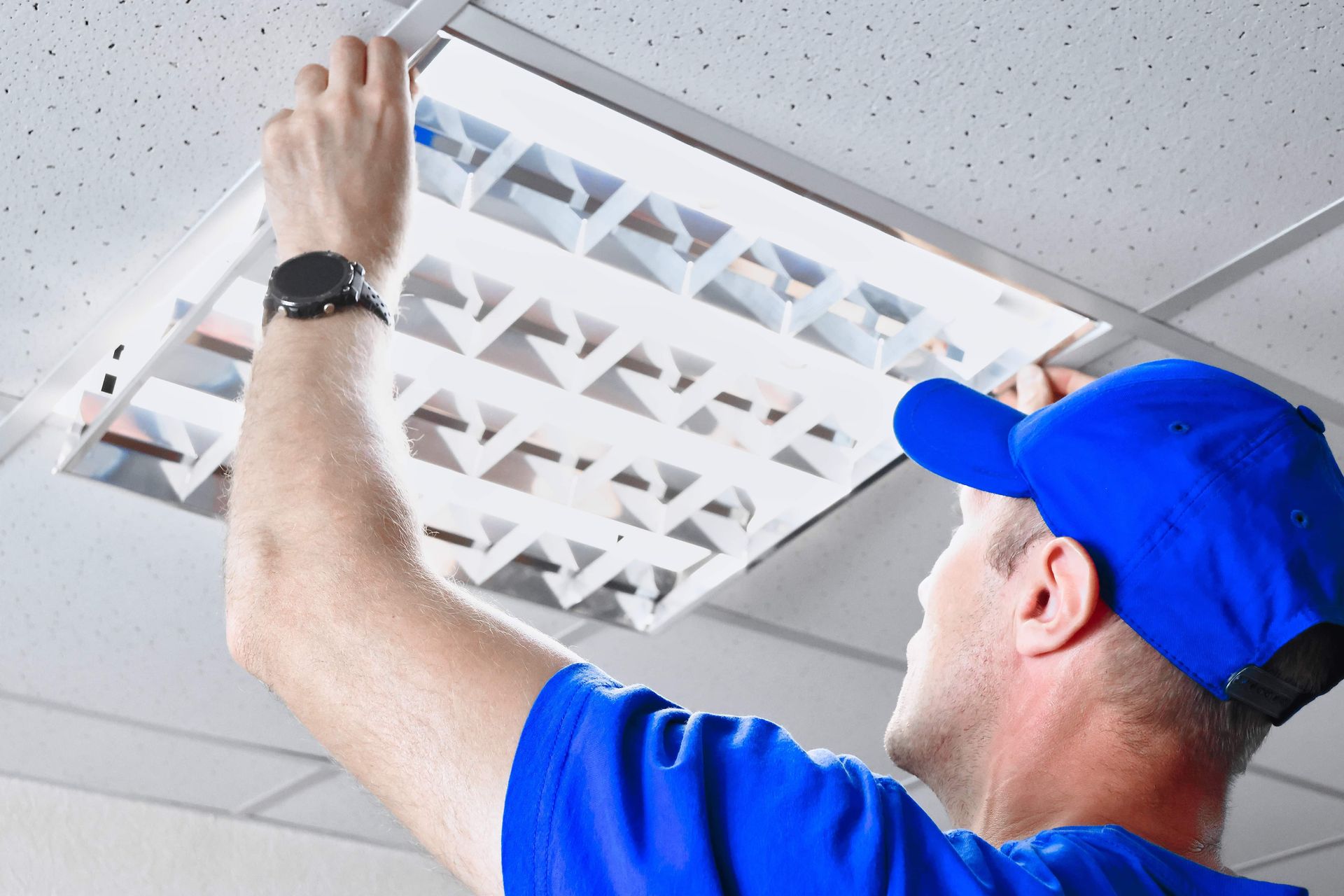 A person wearing a blue shirt and cap installs a square, white-louvered fluorescent light fixture into a drop ceiling.