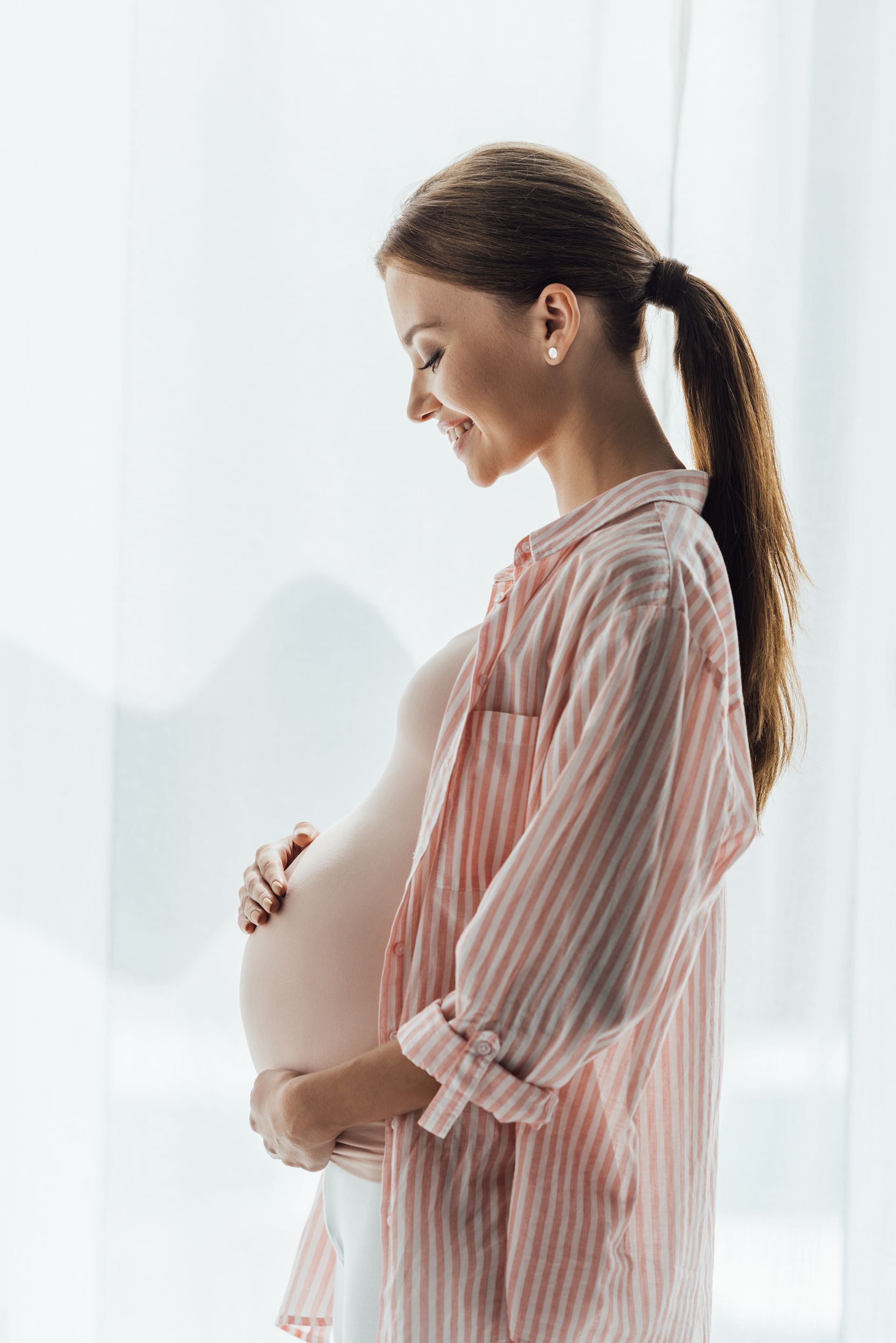 A pregnant woman is standing in front of a window holding her belly.